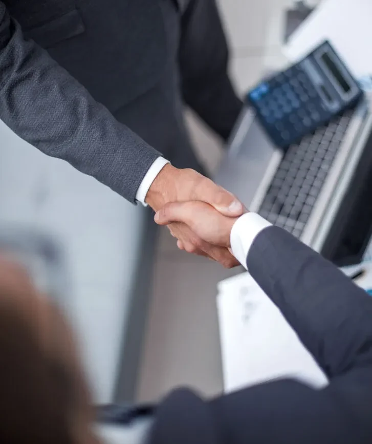 Two businessmen in suits shake hands over a desk with a laptop and documents, symbolizing a successful agreement or partnership Two businessmen in suits shake hands over a desk with a laptop and documents, symbolizing a successful agreement or partnership