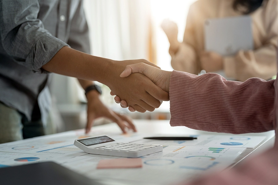 Two people shake hands over a document with charts and a calculator, while a third person observes, indicating a business agreement