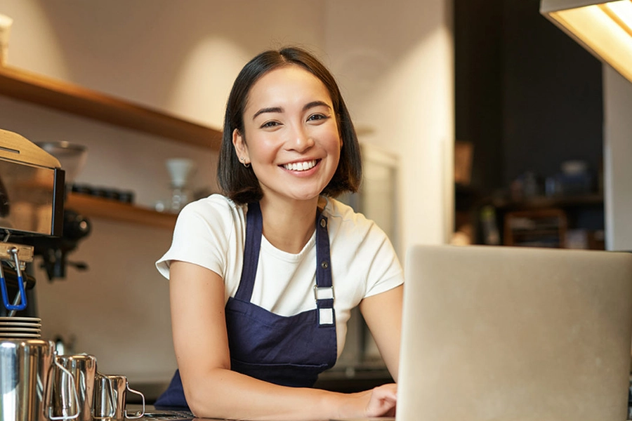 A barista in an apron stands at a counter, working on a laptop with coffee equipment in the background