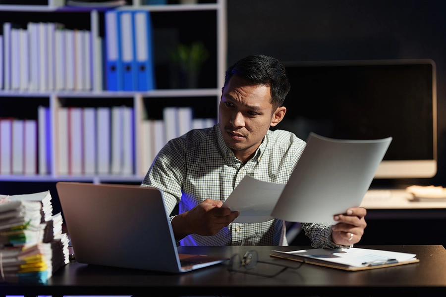 A person in a checked shirt reviews documents while sitting at a desk with a laptop and stacks of papers, in a dimly lit office